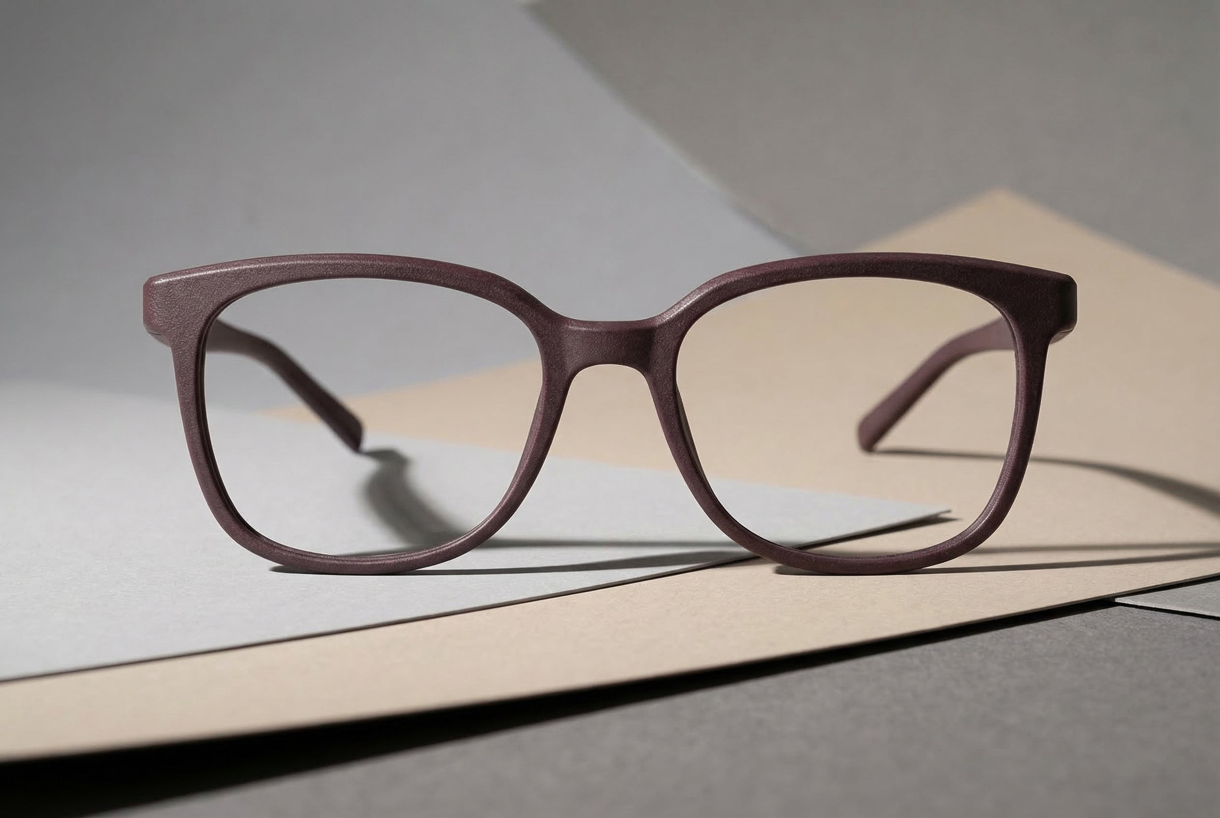 Braun-graue SLS-3D-gedruckte Brille auf gestapelten Papierflächen, leicht schräge Perspektive, weiches Studio-Licht
